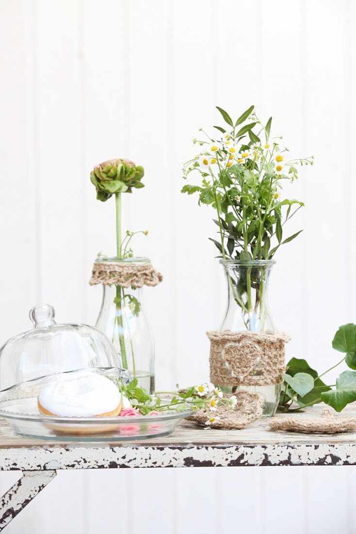 Flowers in carafes decorated with hand-crocheted jute trim and donuts on plate with glass cover