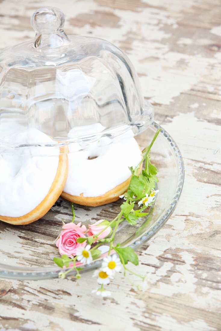 Mit weißem Zuckerguss verzierte Donuts und Blütendeko auf Glasteller mit Glocke