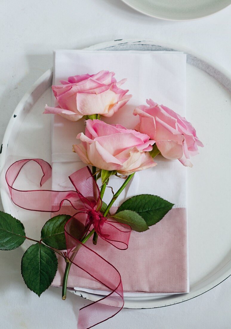 Drei rosa Rosen mit Schleife auf Stoffserviette auf einem Platzteller