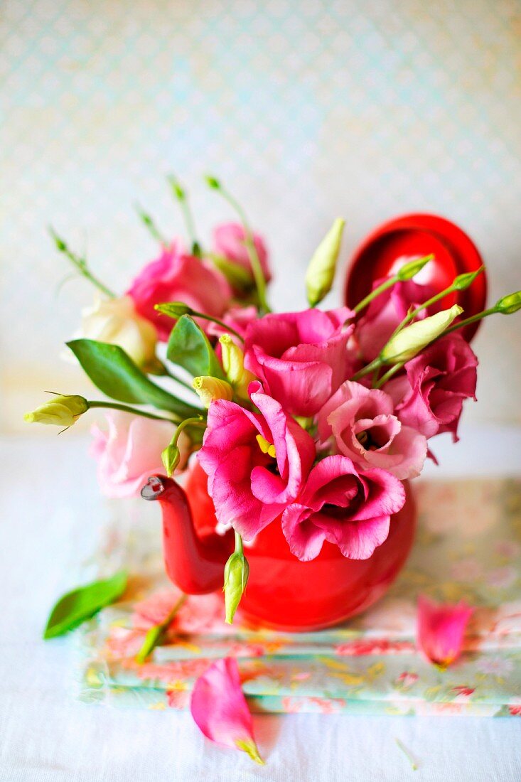 Zartes, kleines Blümensträußchen in rosa gehalten in einem roten Teekännchen arrangiert; einzelne Blütenblättchen liegen auf den gefalteten, blumigen Servietten auf dem weißen Untergrund