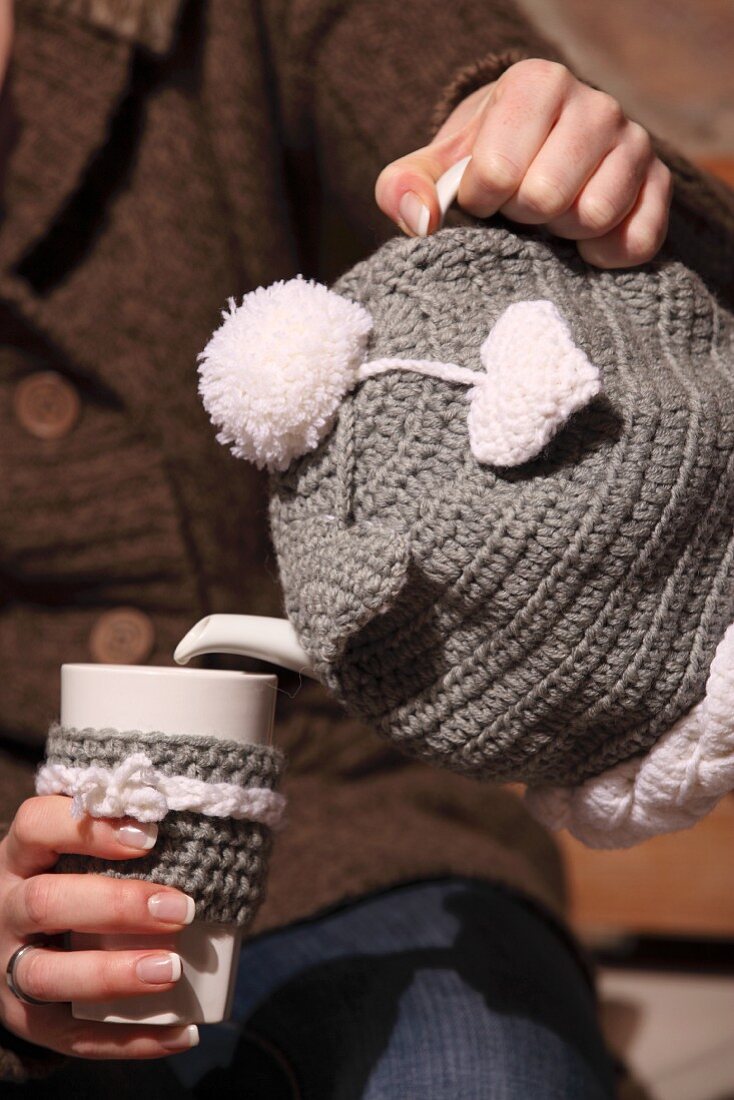Woman pouring tea - hand-crocheted tea cosy on teapot and cover on beaker held in other hand