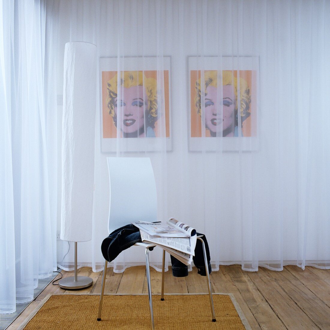 White shell chair in front of standard lamp next to transparent curtains; pair of portraits of Marilyn Monroe behind curtains