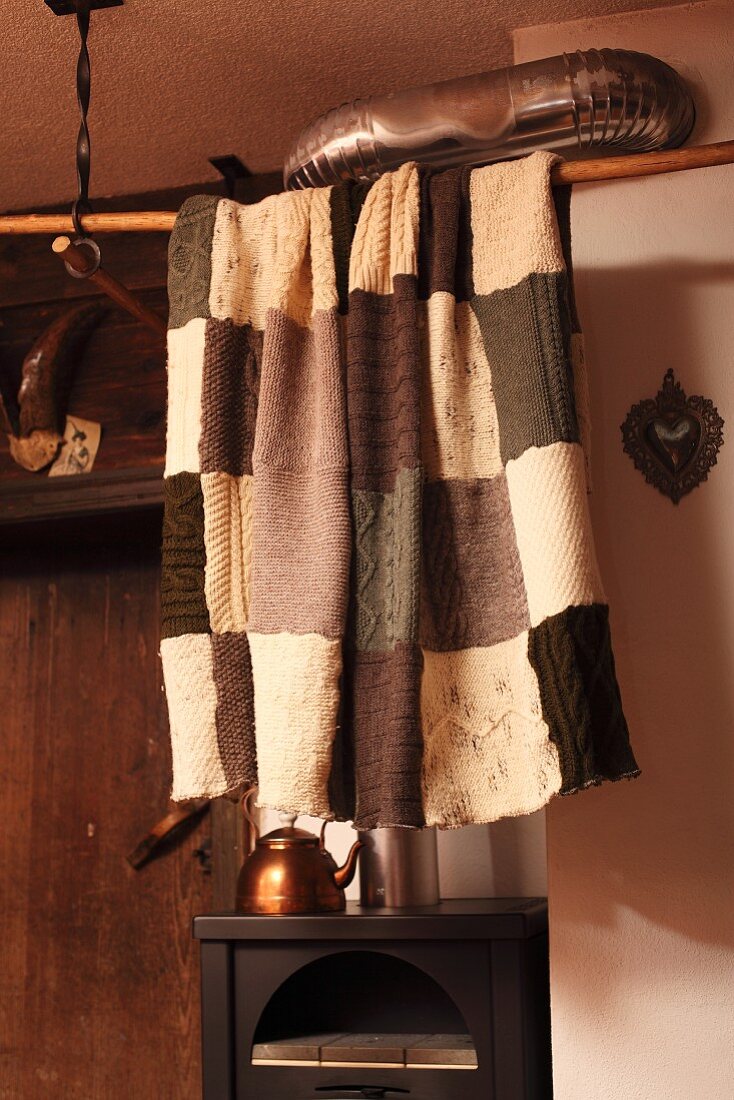 Patchwork blanket hanging from wooden rod below ceiling in front of copper tea kettle on wood-burning stove