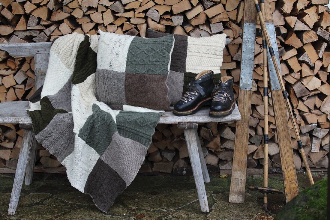 Vintage skiing equipment, knitted patchwork blanket and cushions on weathered wooden bench in front of stacked firewood