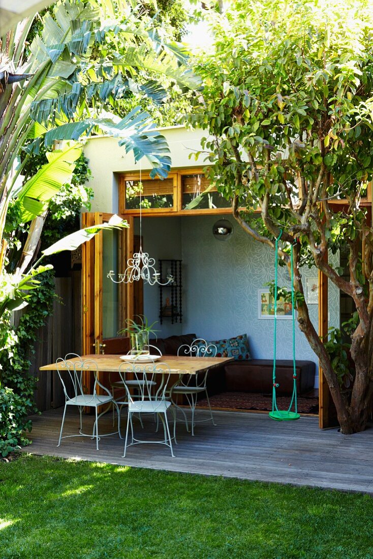 Tropischer Garten mit Tisch und Stühlen auf kleinem Holzdeck vor offener Terrassentür und Blick in ein Zimmer mit Liege