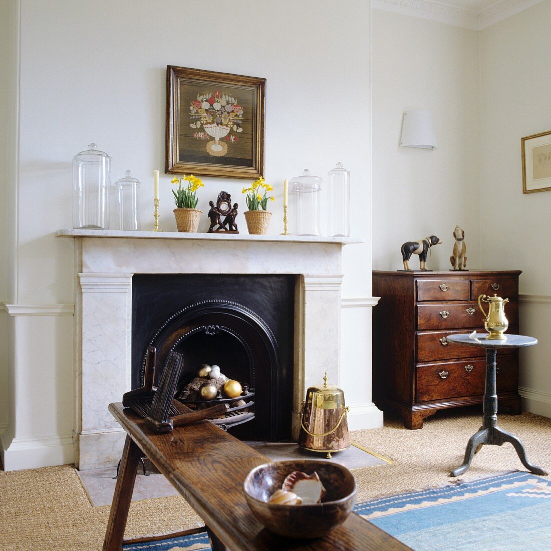 Interior with wooden bench in front of open fireplace, glass vessels on mantelpiece and antique, wooden chest of drawers in corner