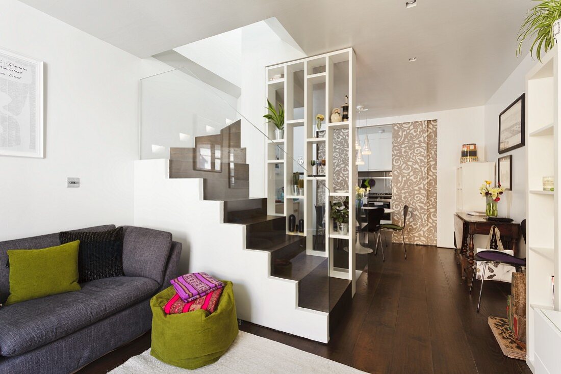 Offener Wohnraum mit hoher Regalwand als Raumteiler und Blick auf dunkelbraune Treppe mit Glasgeländer; davor eine graue Couch und ein grüner Sitzsack