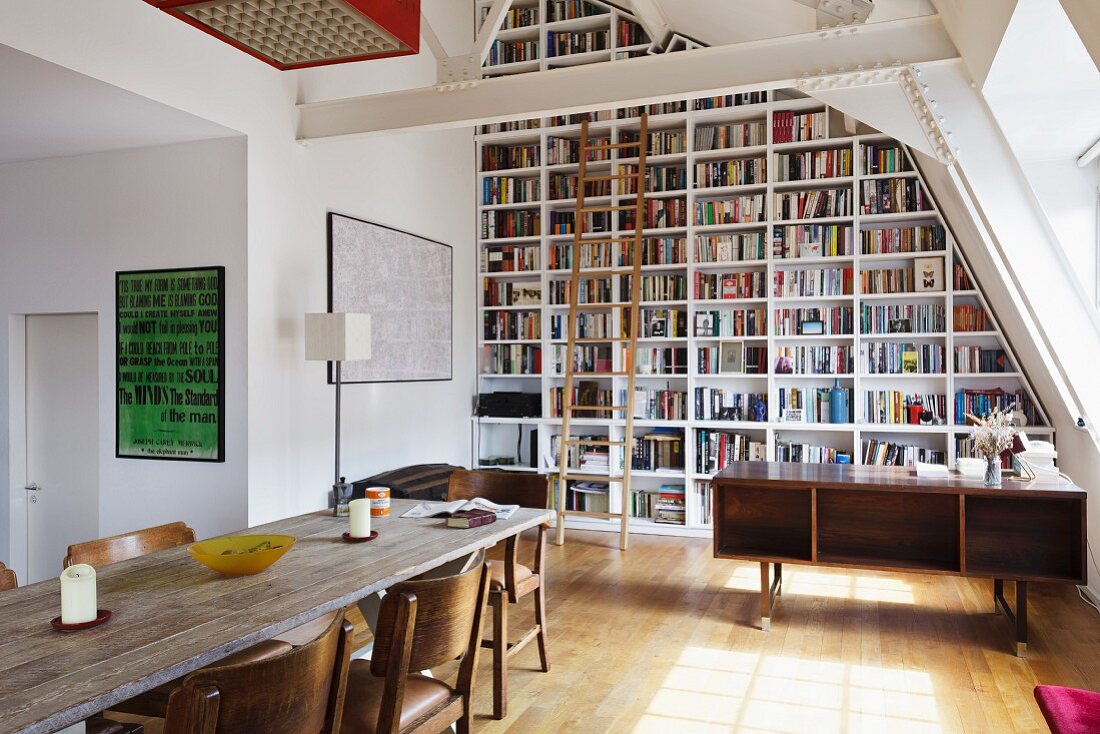 Raumhohe Bücherwand mit davorstehender Holzleiter unter der Dachschräge; im Vordergrund eine lange Holztafel und gepolsterte Stühle