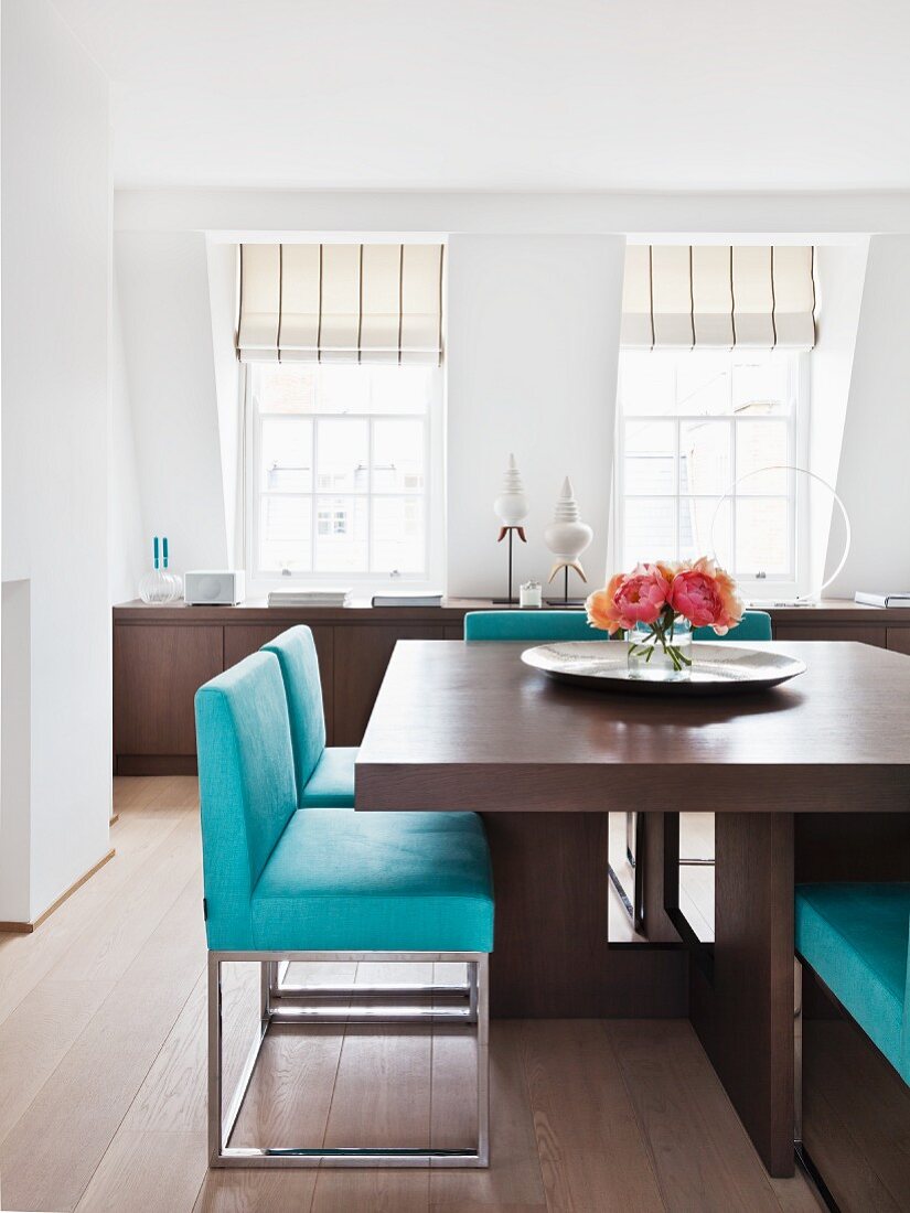 Brown, solid wood dining table with turquoise metal chairs; long sideboard below windows in background