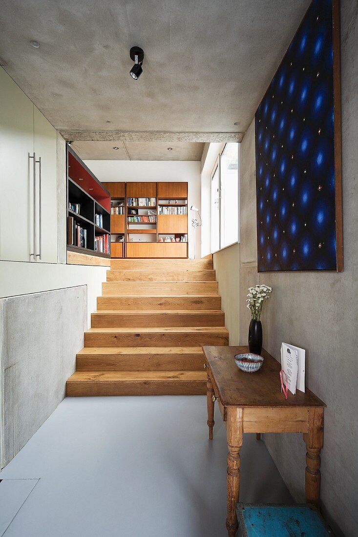 Vintage Wandtisch und moderne Kunst an Betonwand; im Hintergrund eine helle Holztreppe und ein großer Bücherschrank