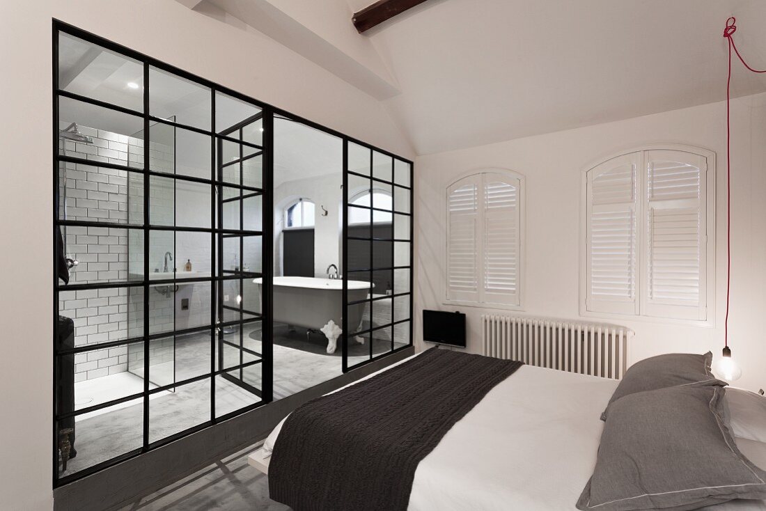 Minimalist bedroom in black, grey and white; glass partition offers view into bathroom with antique, free-standing bathtub