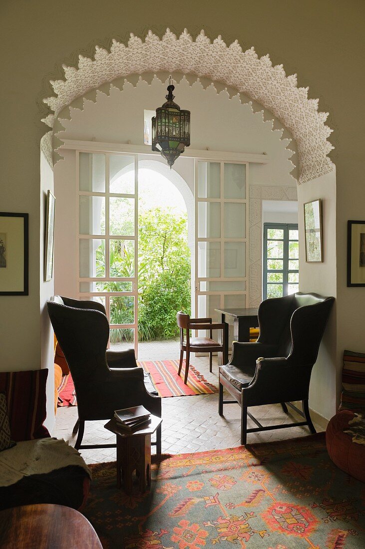 Two wingback armchairs under Moroccan-style archway with ornate details; view into green courtyard in background