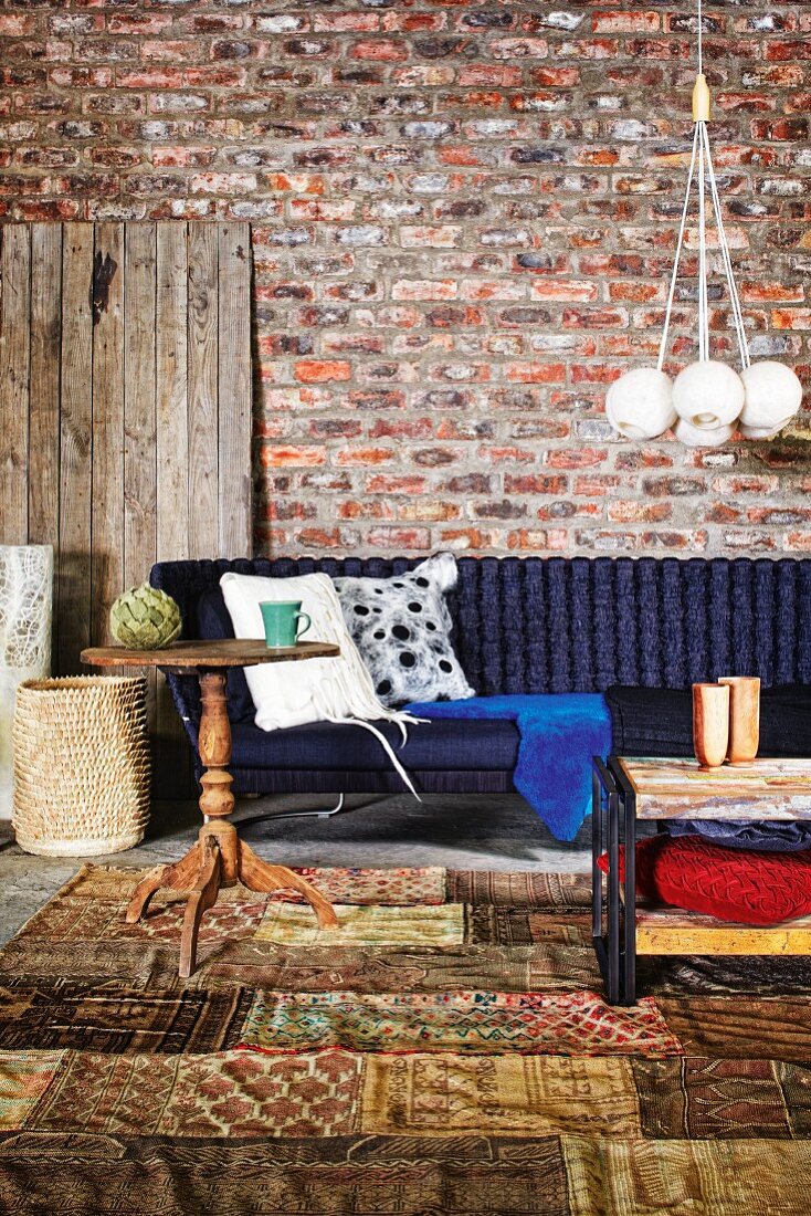 Rustikaler Holz Beistelltisch auf Patchwork Teppich vor dunkelblauem Sofa an rustikaler Ziegelwand; Hängeleuchte mit gebündelten, kugelförmigen Schirmen