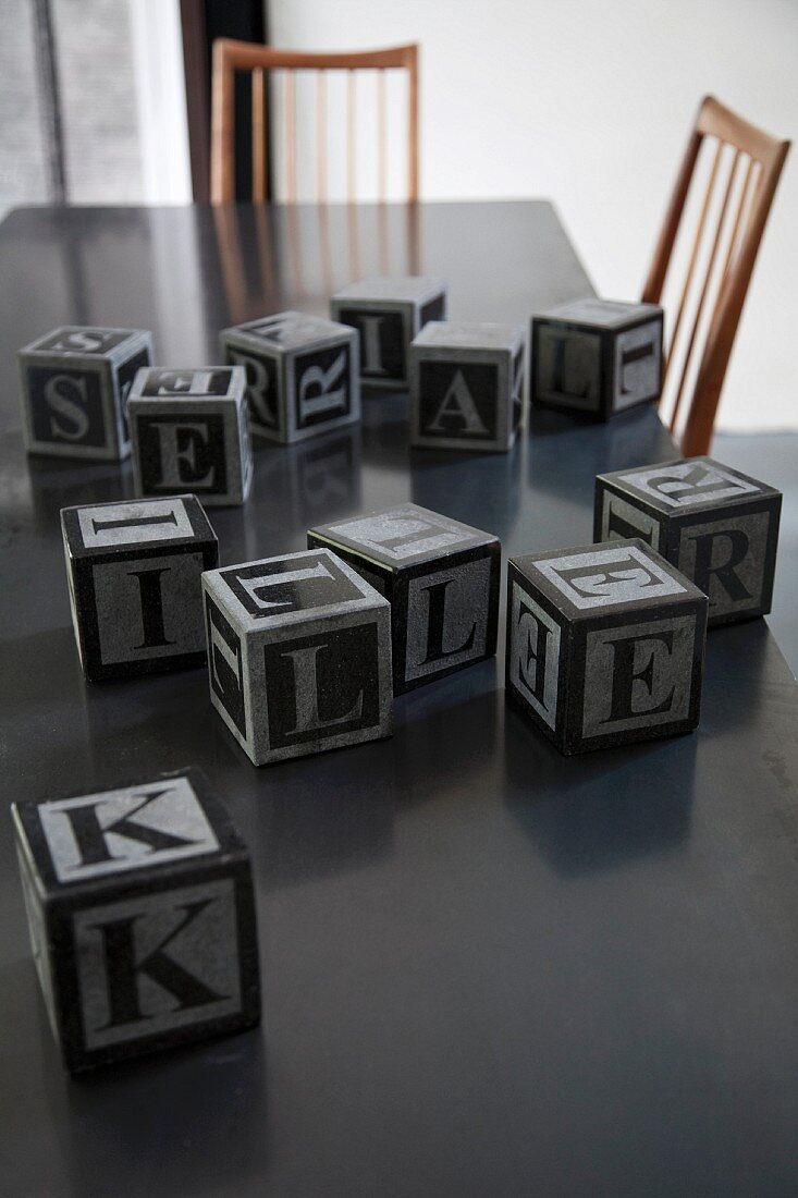 Mehrere Buchstabenwürfel auf dunkler Tischplatte mit Stühlen im Fifitiesstil