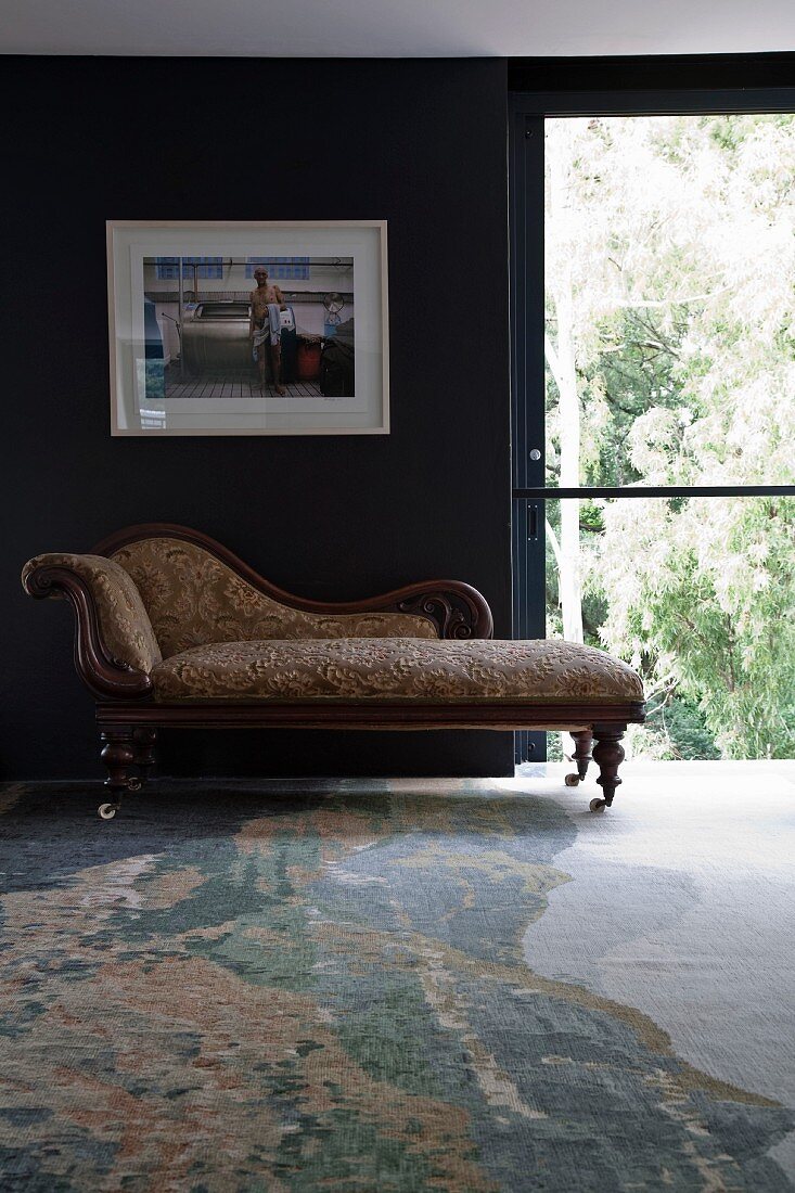 Antike Chaise Longue auf Rollen vor dunkel getönter Wand mit gerahmter Fotografie neben Schiebefenstertür; gemusterter Teppich mit Natursteinoptik