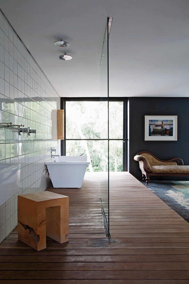 Bedroom with open-plan bathroom on wooden platform; bathtub, wooden stool and rain shower behind glass screen, large window and antique recamier in background