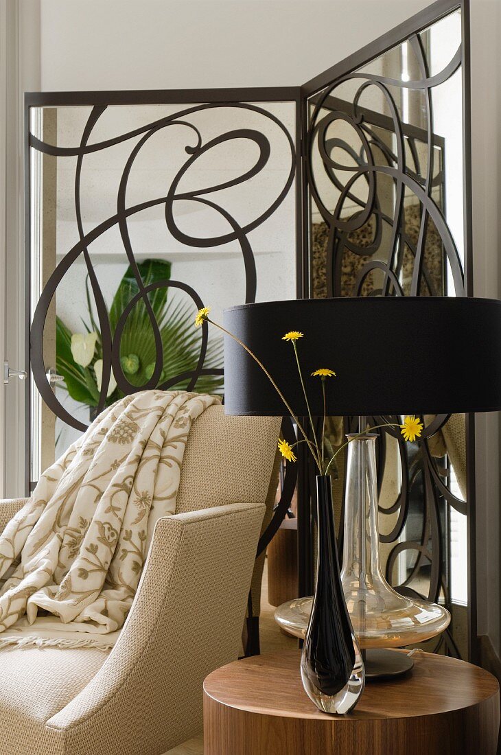 Black glass vase of flowers and table lamp with black lampshade on side table in front of pale armchair and modern, wrought iron screen