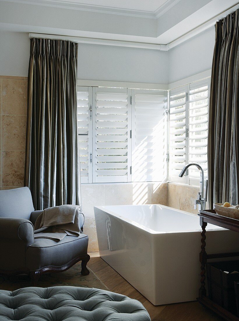 Upholstered armchair next to free-standing bathtub below windows with floor-length curtains