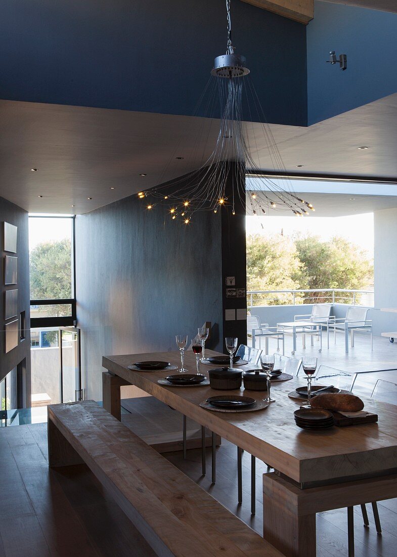 Gedeckter Esstisch mit Holzbank unter moderner Hängeleuchte in offenem, minimalistischem Wohnraum
