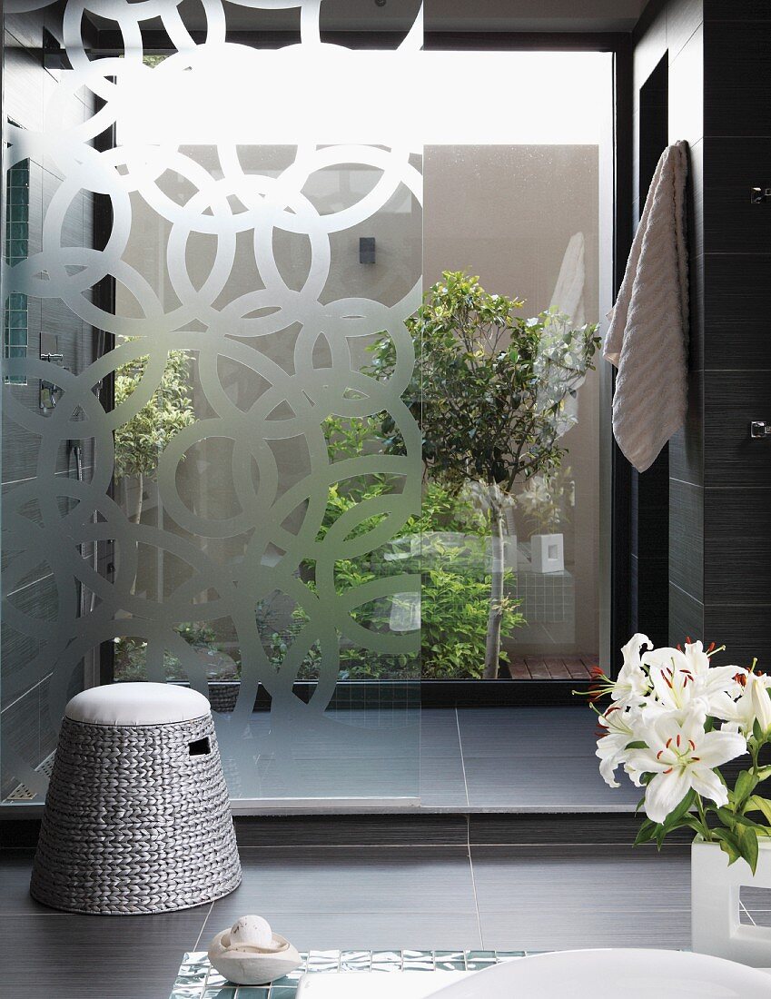 Bathroom with dark grey tiles and shower area in front of glass wall with view into planted courtyard