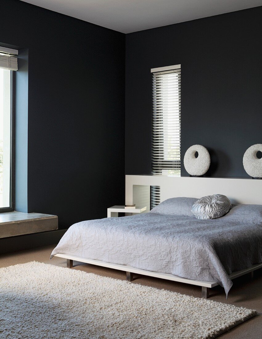 Doppelbett mit hellgrauer Tagesdecke, vorne heller Teppich in schwarz getöntem, minimalistischem Schlafzimmer