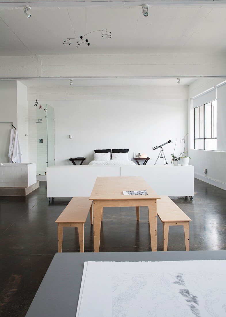 Tisch und Bänke aus hellem Holz auf grauem Estrichboden vor weiss gehaltenem Schlafbereich in loftartigem Raum