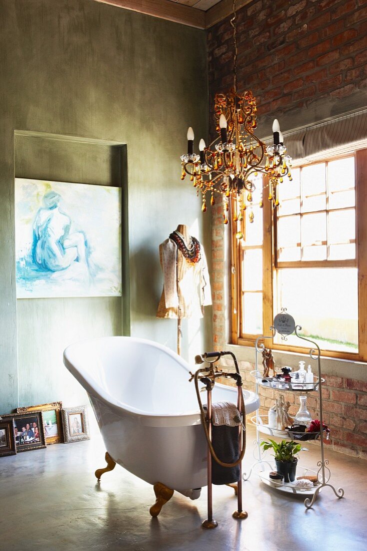 Free-standing vintage bathtub below chandelier in rustic bathroom with concrete floor