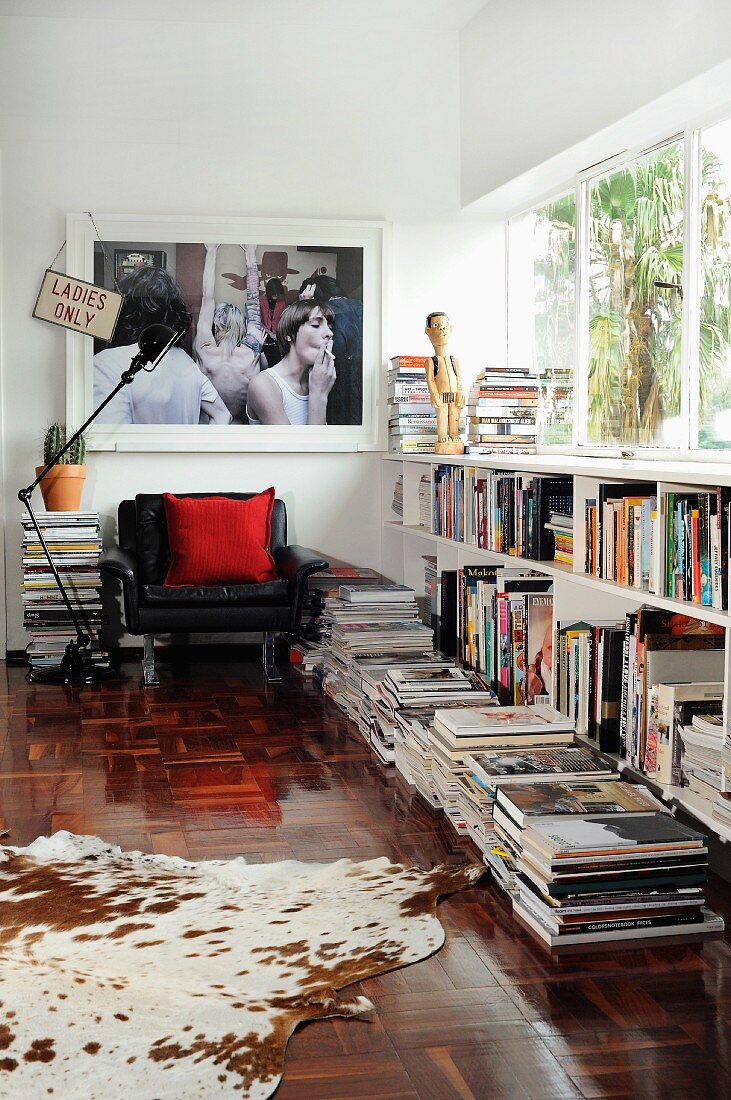Cowhide rug and stacked books on floor next to half-height shelving below ribbon window; armchair & large photo in background