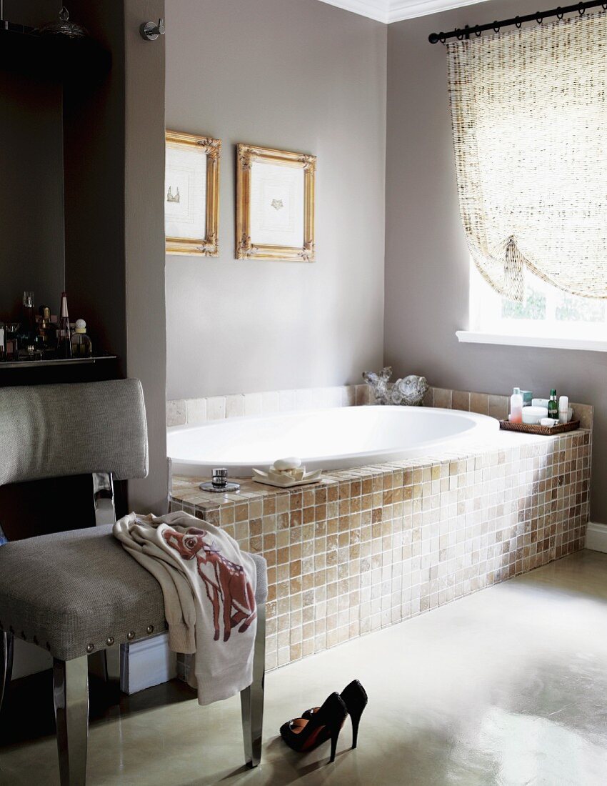 Gepolsterter Stuhl neben Badewanne mit gefliester Seitenwand in wohnlichem Badezimmer