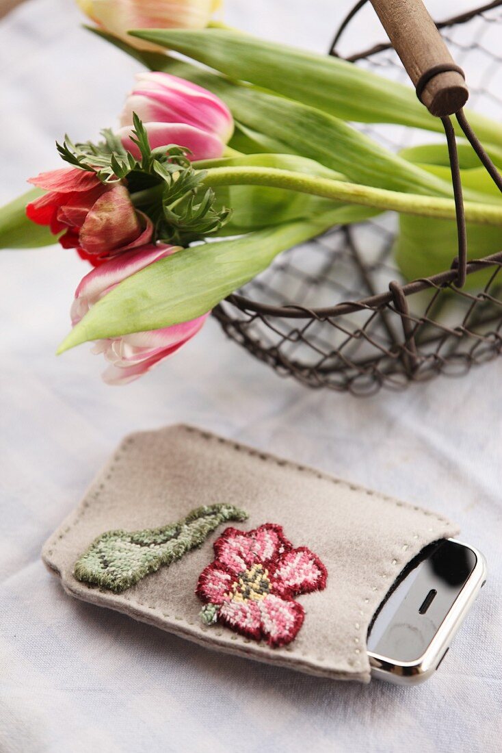 Handy in handbestickter Tasche neben Drahtkorb mit Frühlingsblumen