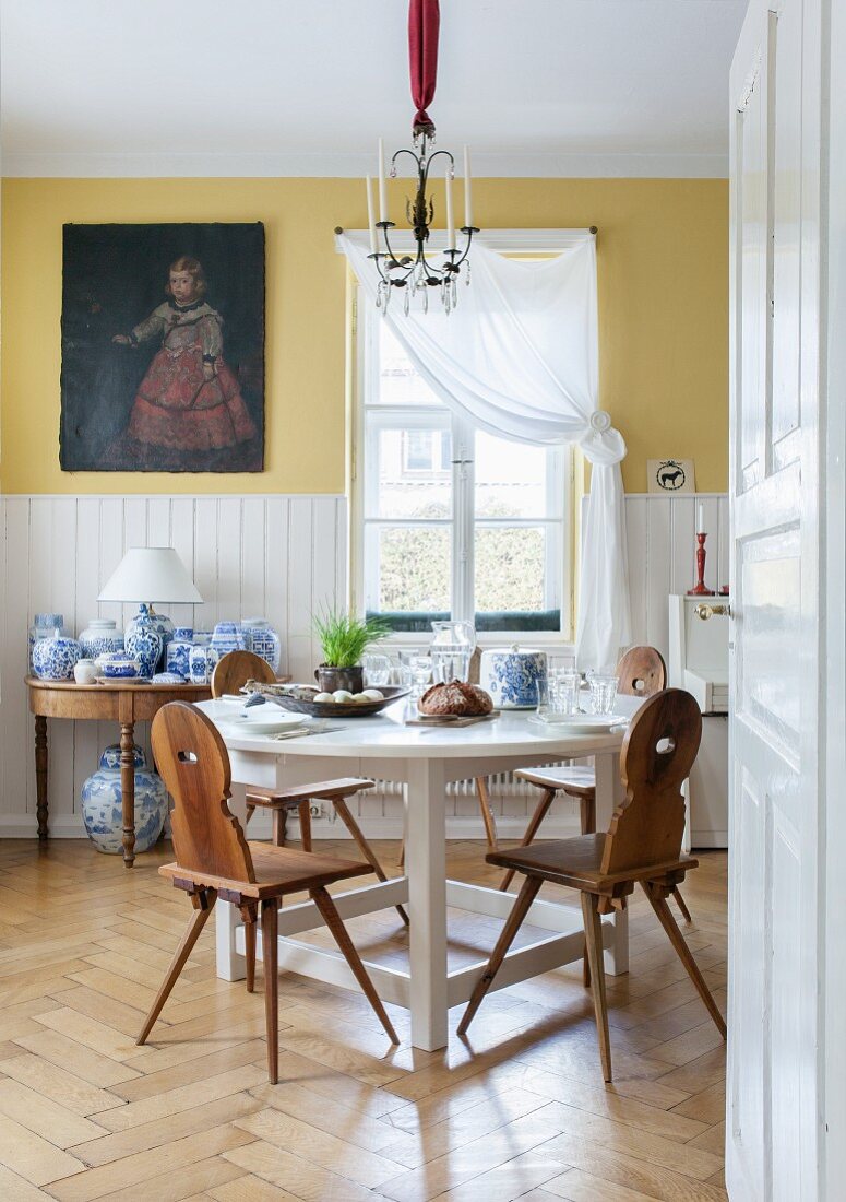 Traditionelle, bayerische Brettstühle an weißem, rundem Esstisch und Sammlung blauweisser Porzellangefässe auf Konsolentisch unter altem Ölgemälde