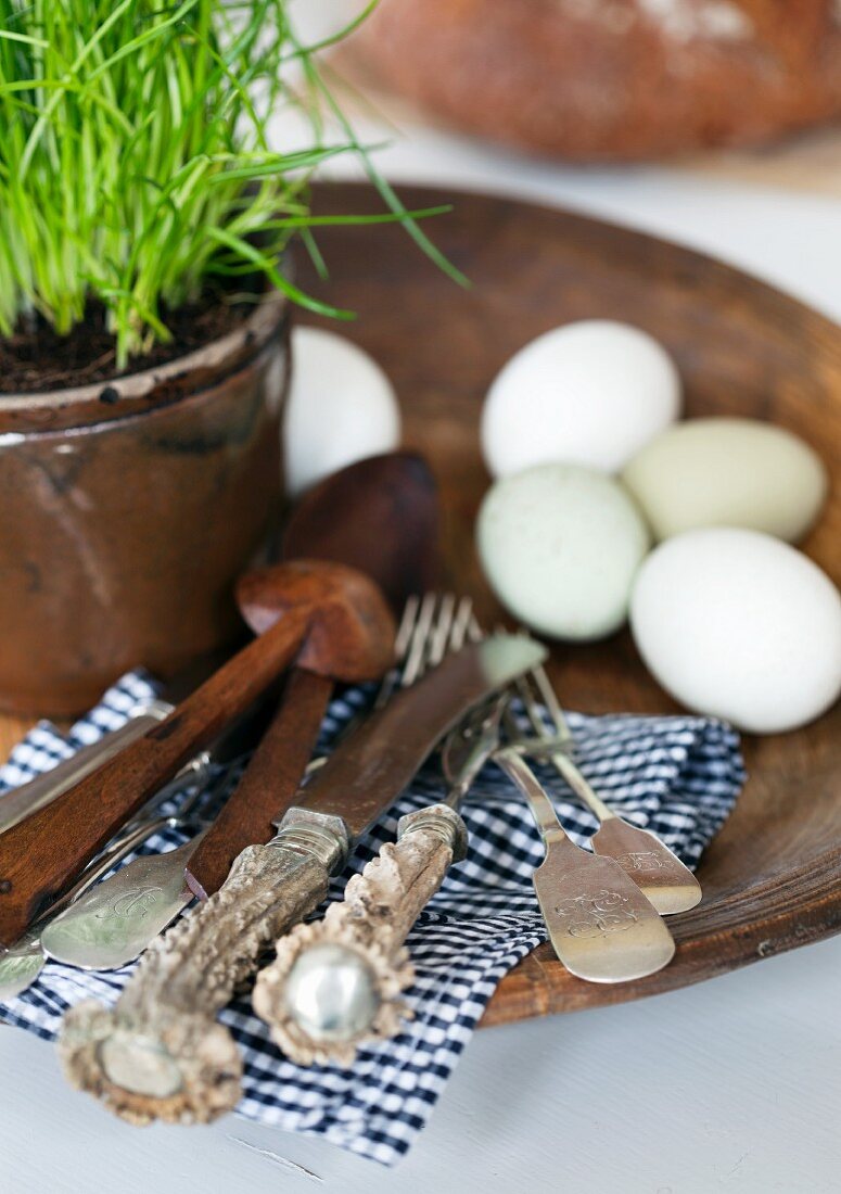 Handgeschnitztes Salatbesteck und altes Silberbesteck mit Spaten und Horngriffen auf schwarzweisser Karoserviette; Eier und Schnittlauchtopf auf Steingutteller