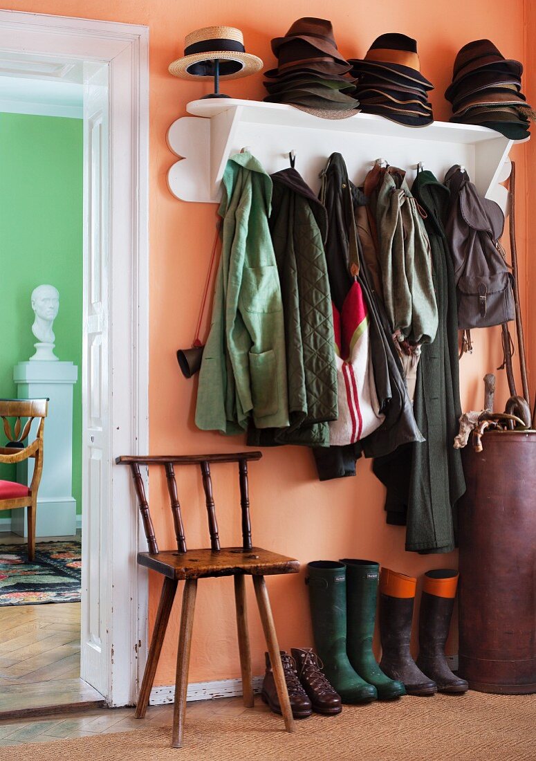 Garderobe mit Hutsammlung, Schirmtonne und Holzstuhl neben Gummistiefel in lachsfarbener Diele; seitlich Blick auf Männerbüste vor lindgrüner Wand im Wohnzimmer