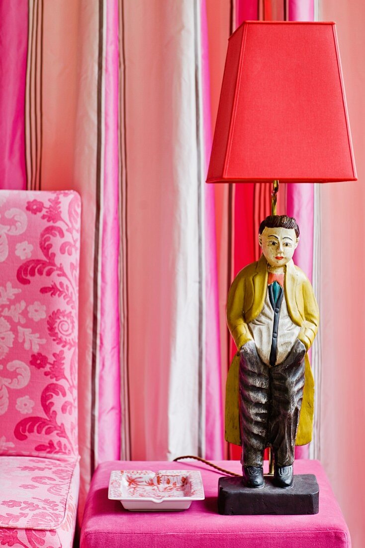 Als Lampe gestaltete Chinesenfigur auf pinkfarbenem Samtpouf, ergänzt mit gestreiften Seidentaftvorhängen und bedruckter Leinenhusse
