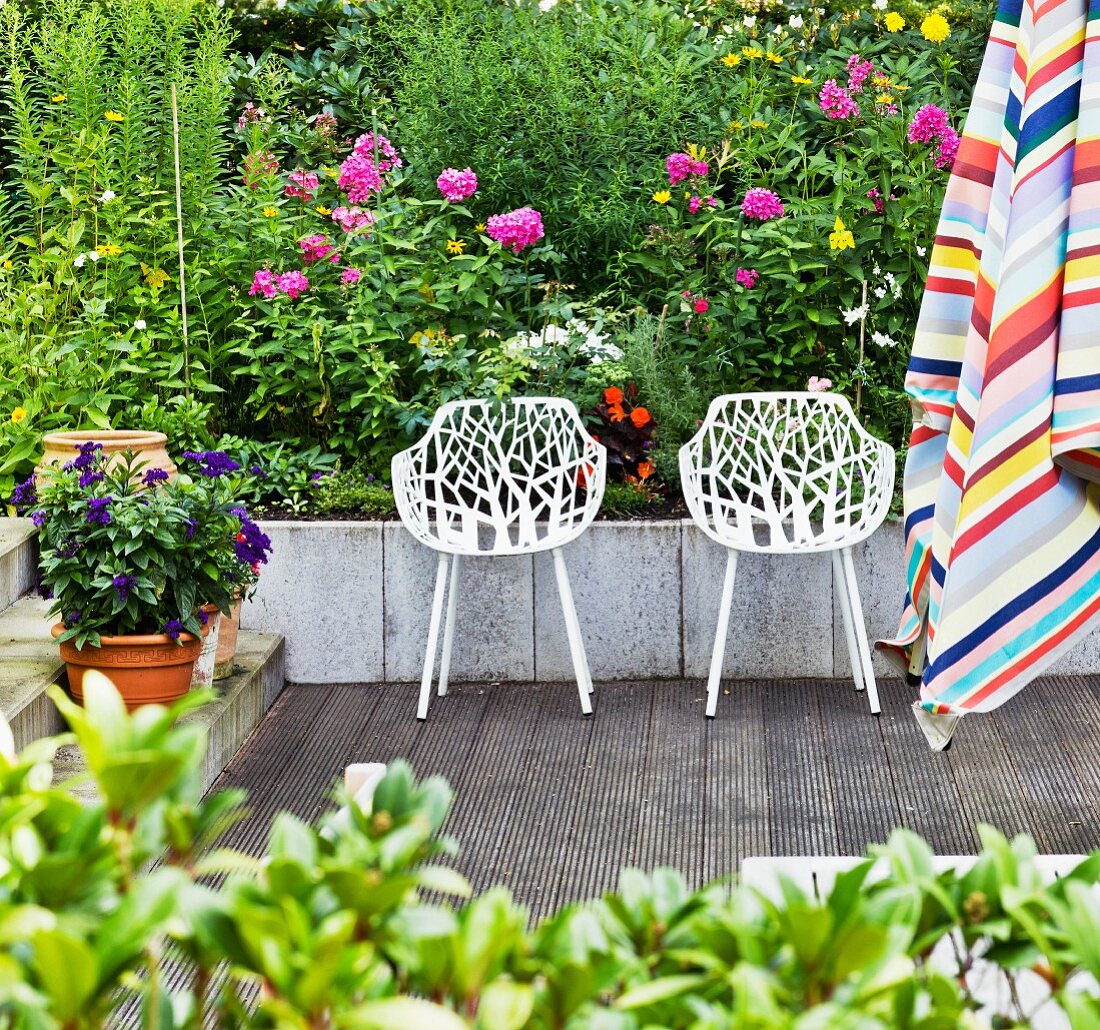 Durchbrochene, weiße Schalensessel auf Terrasse vor Blumenbeet; im Vordergrund gestreifter Sonnenschirm