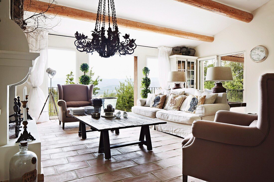 Armchairs and sofa around rustic wooden table on terracotta floor in country-style living room
