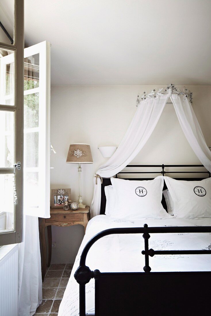 Double bed with black metal frame and canopy of white, airy fabric in country-style interior