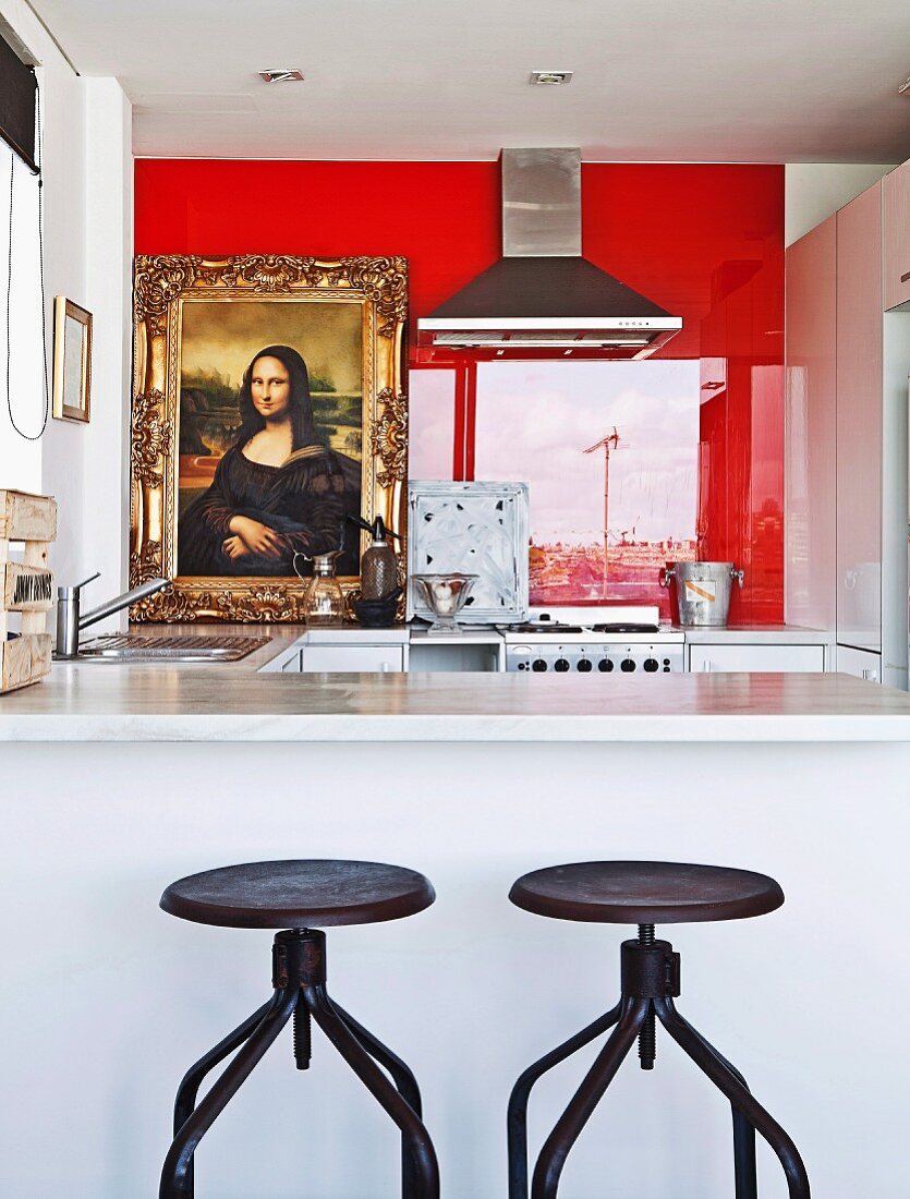Vintage swivel stools with metal bases at white counter in open-plan kitchen and view of Mona Lisa reproduction against red glass wall