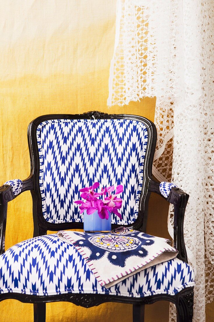 Gepolsterter Stuhl in postmodernem Stil mit weiss blauem Bezug und schwarzem Holzgestell vor gelber Wand mit Spitzenstoff drapiert