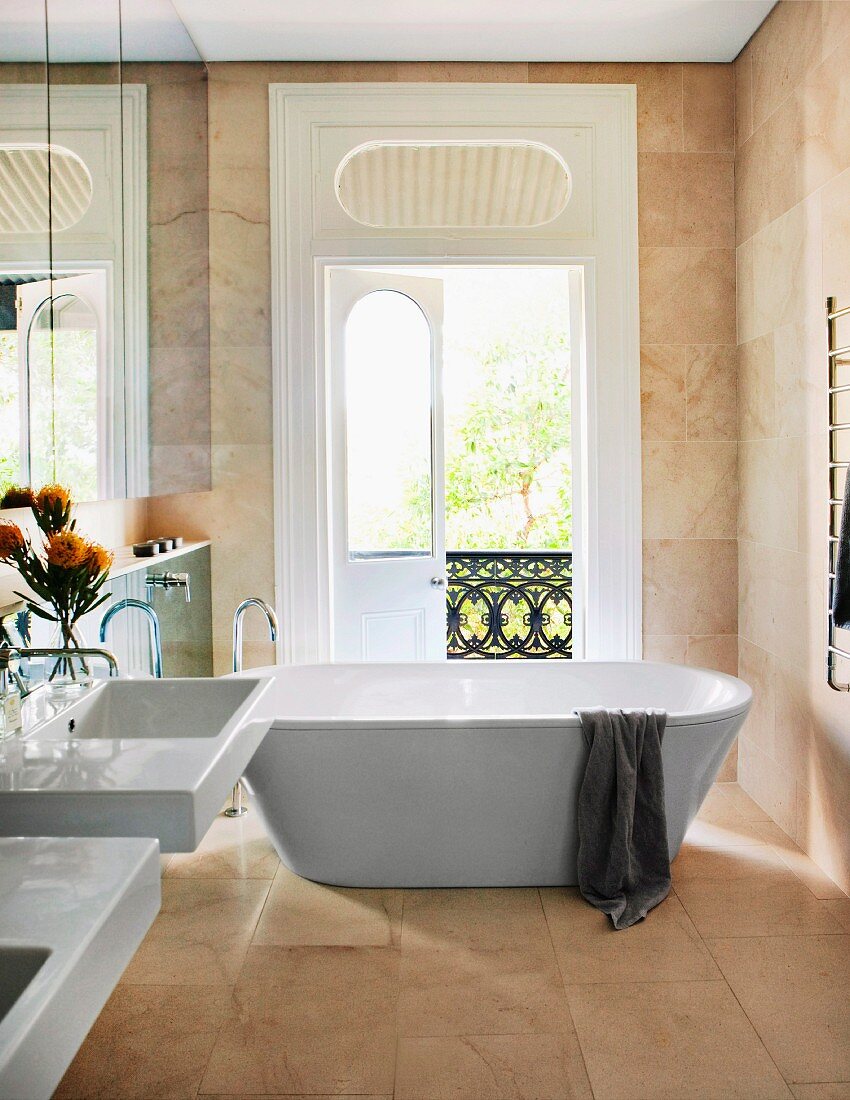 Free-standing bathtub in front of open balcony door and mirrored cabinet doors above sinks in elegant, marble bathroom