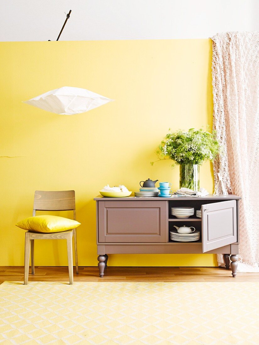 Kleine Landhaus-Kommode für Geschirr, moderner Holzstuhl und japanische Reispapierlampe vor gelber Wand