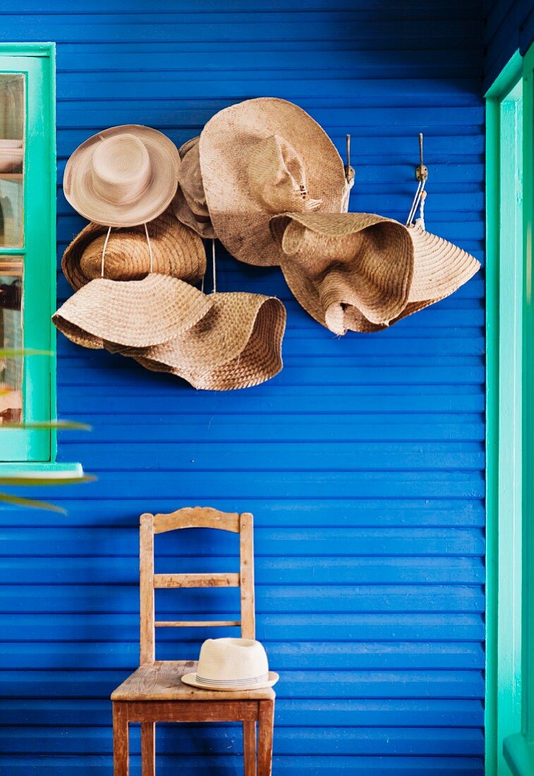 Garderobenhaken mit Strohhüten und Stuhl an blauer Wand mit türkisfarbenen Fensterrahmen