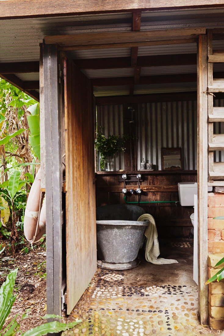 Outdoor bathroom with zinc bathtub in wooden structure with corrugated metal cladding and pebble mosaic floor