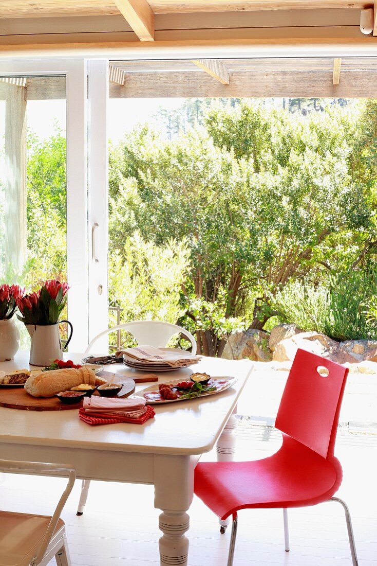 Roter Stuhl an rustikalem Holztisch mit angerichteten Vorspeisen; Blick durch Terrassenfenster in sonnenbeschienenen Garten