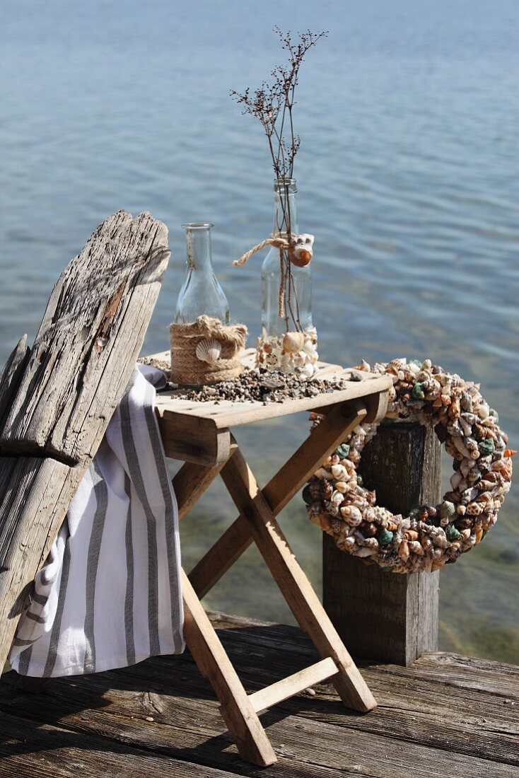 Bottle decorated with shells and jute rope braid on folding table and wreath of shells on wooden jetty next to lake