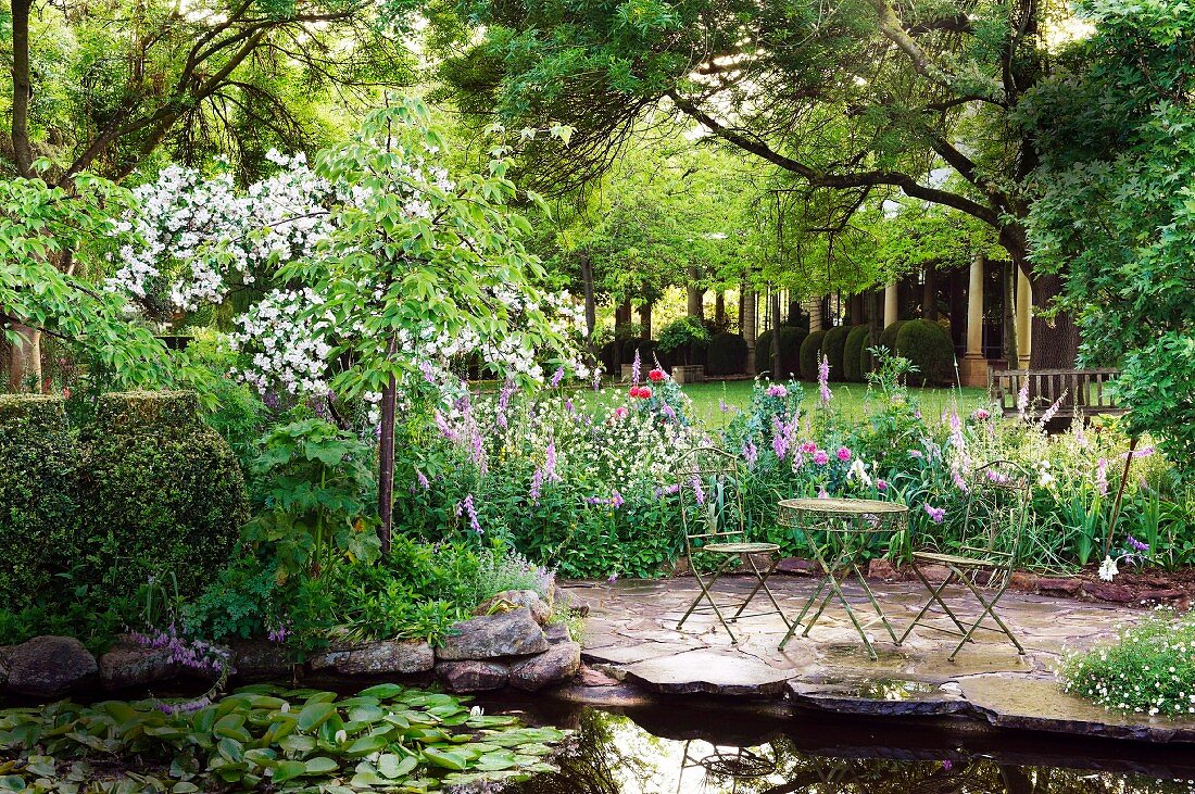 Von blühenden Stauden begrenzter, plattenbelegter Sitzplatz an Seerosenteich in weitläufigem Garten
