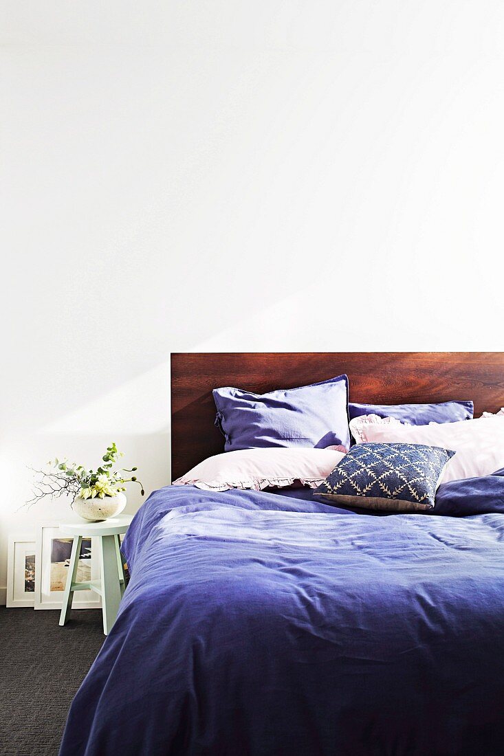 Doppelbett mit Kopfteil aus rötlichem Edelholz und lila Bettwäsche, daneben Vintage Hocker mit kleiner Blumenvase