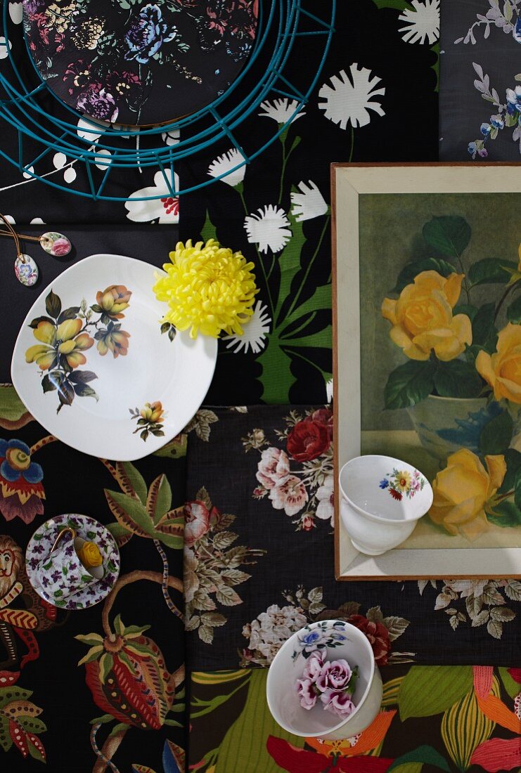 Blick von oben auf Tassen und Teller mit gelber Blüte und gerahmtes Bild auf Stoffen mit Blumenmotiven