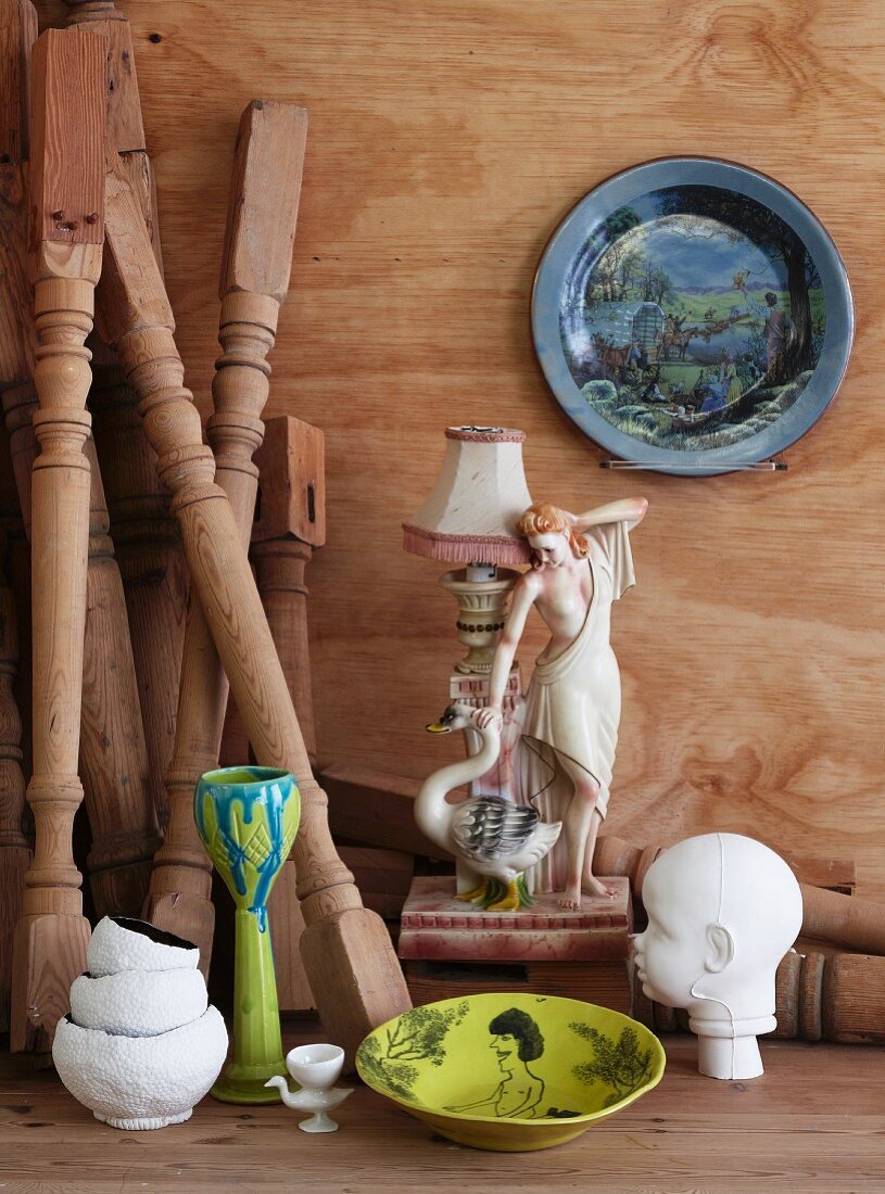 Kleine Tischlampe aus Porzellan mit Frauenfigur, bemalte Teller und Kinderkopfskulptur; in der Ecke lehnende gedrechselte Holzbeine
