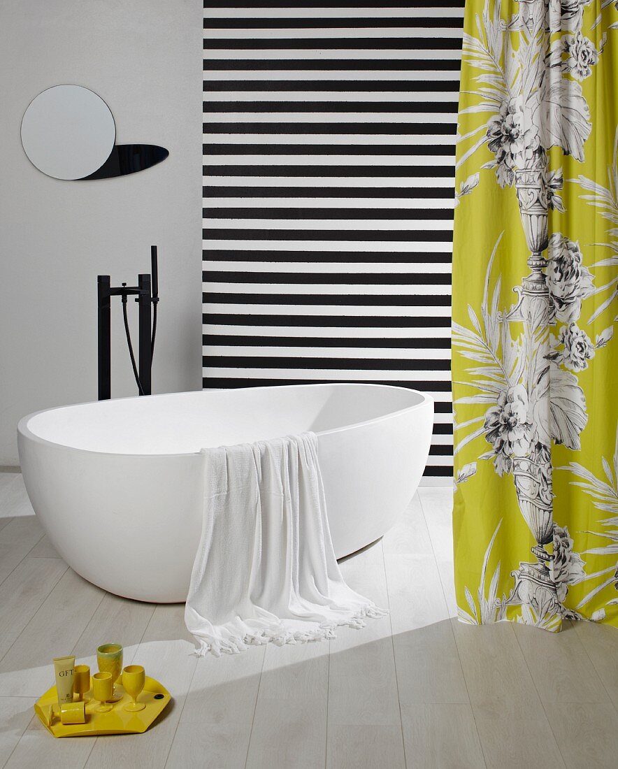 Towel hanging over edge of free-standing white bathtub amongst hanging lengths of fabric and small, round mirror on wall